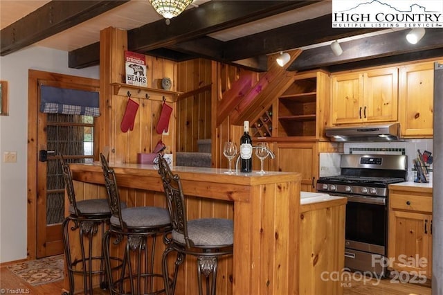 kitchen featuring under cabinet range hood, stainless steel gas range oven, open shelves, and beamed ceiling