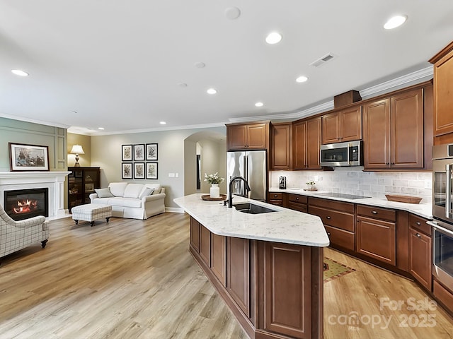 kitchen featuring light stone counters, light wood-type flooring, an island with sink, stainless steel appliances, and decorative backsplash