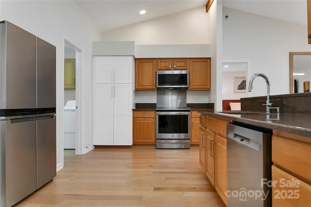 kitchen featuring appliances with stainless steel finishes, a sink, washer / dryer, light wood finished floors, and high vaulted ceiling