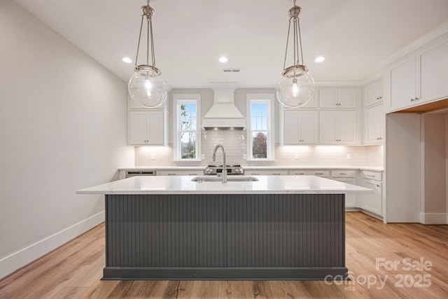 kitchen with backsplash, premium range hood, light countertops, an island with sink, and white cabinetry