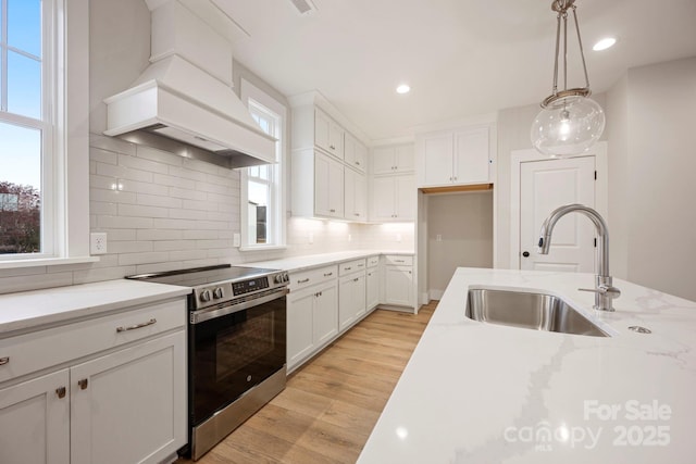 kitchen with light wood finished floors, backsplash, premium range hood, stainless steel electric stove, and a sink