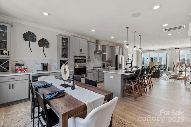 kitchen featuring stainless steel appliances, decorative light fixtures, an island with sink, wall chimney range hood, and gray cabinetry