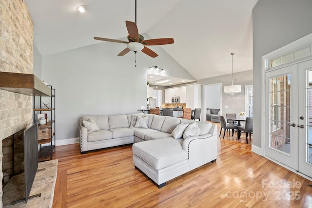 living room with ceiling fan, light hardwood / wood-style floors, a stone fireplace, high vaulted ceiling, and french doors