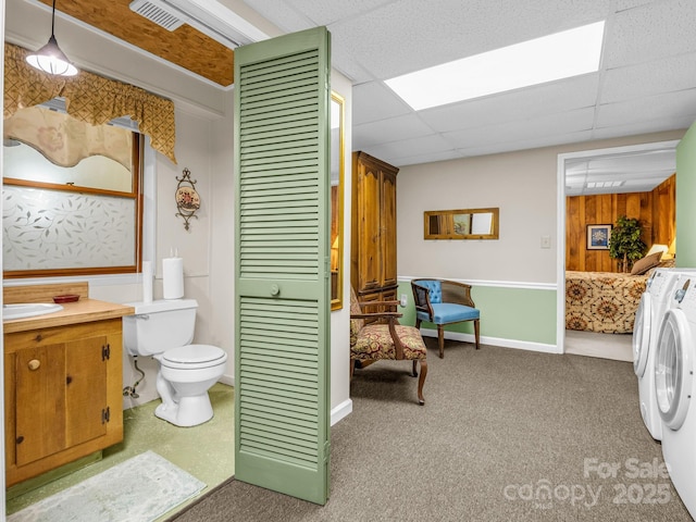 bathroom featuring wooden walls, vanity, toilet, washing machine and dryer, and a drop ceiling