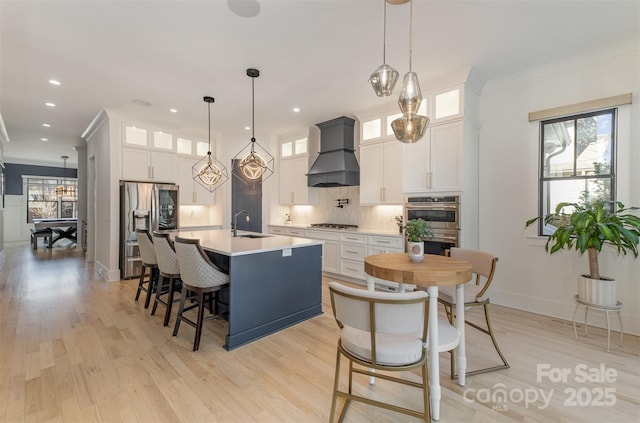 kitchen with decorative light fixtures, a kitchen island with sink, white cabinetry, and custom range hood