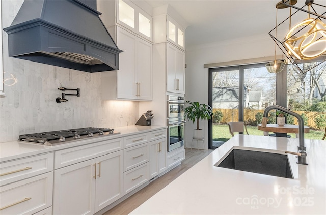 kitchen featuring custom exhaust hood, white cabinetry, sink, backsplash, and a notable chandelier