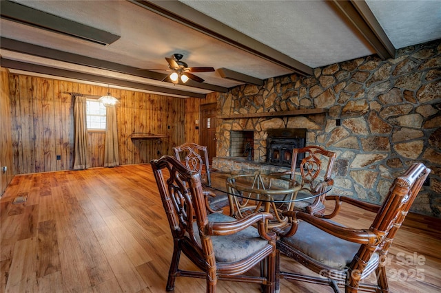 unfurnished dining area with wood walls, wood-type flooring, beamed ceiling, ceiling fan, and a wood stove