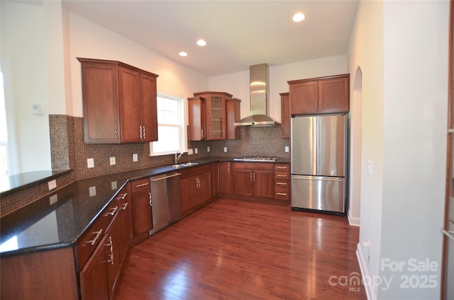 kitchen with stainless steel appliances, wall chimney exhaust hood, backsplash, dark stone counters, and dark wood-type flooring