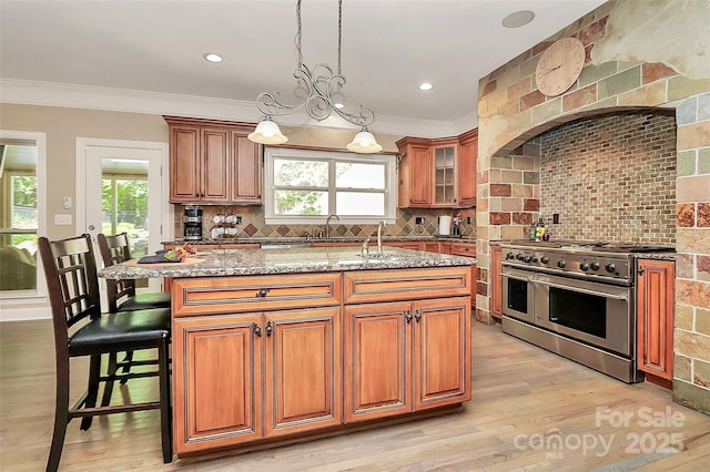 kitchen with a breakfast bar area, light stone countertops, an island with sink, decorative backsplash, and range with two ovens