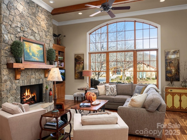 living room with a stone fireplace, ceiling fan, wood-type flooring, ornamental molding, and beamed ceiling