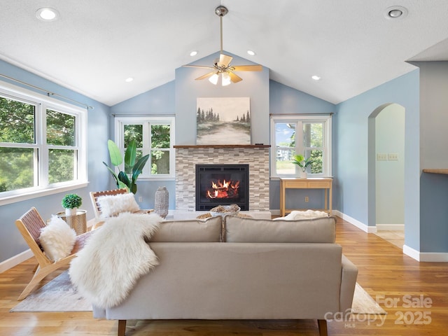 living room with ceiling fan, lofted ceiling, a stone fireplace, and light wood-type flooring