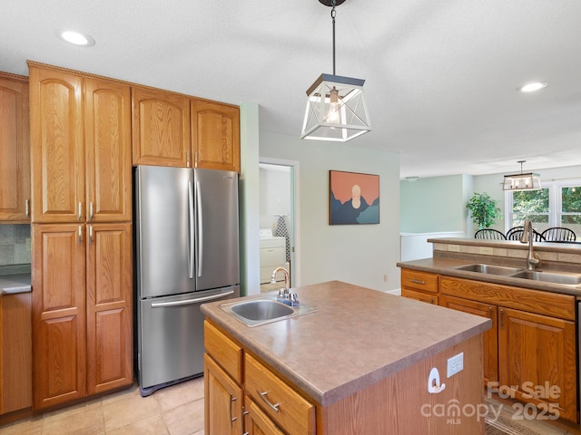 kitchen with sink, a kitchen island with sink, stainless steel refrigerator, and pendant lighting