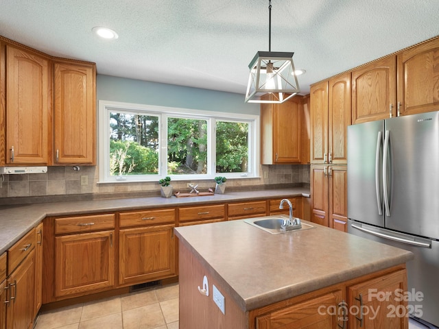 kitchen with pendant lighting, decorative backsplash, sink, stainless steel refrigerator, and a center island with sink