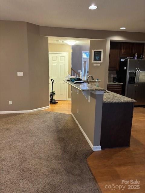 kitchen with a breakfast bar, light stone countertops, an island with sink, stainless steel fridge with ice dispenser, and dark brown cabinets