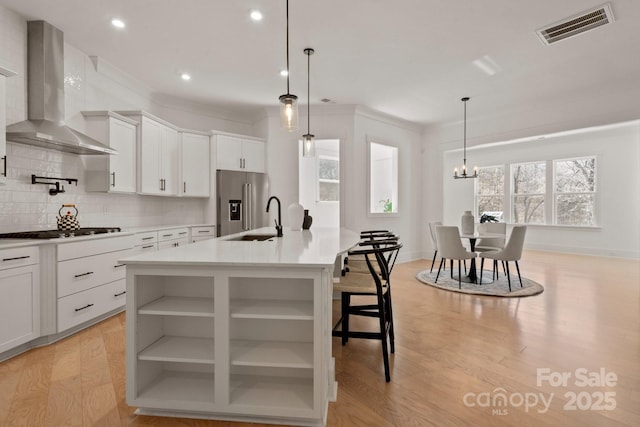 kitchen featuring open shelves, visible vents, a kitchen island with sink, a sink, and wall chimney exhaust hood