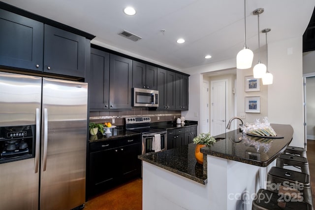 kitchen featuring stainless steel appliances, a kitchen island with sink, hanging light fixtures, and dark stone counters