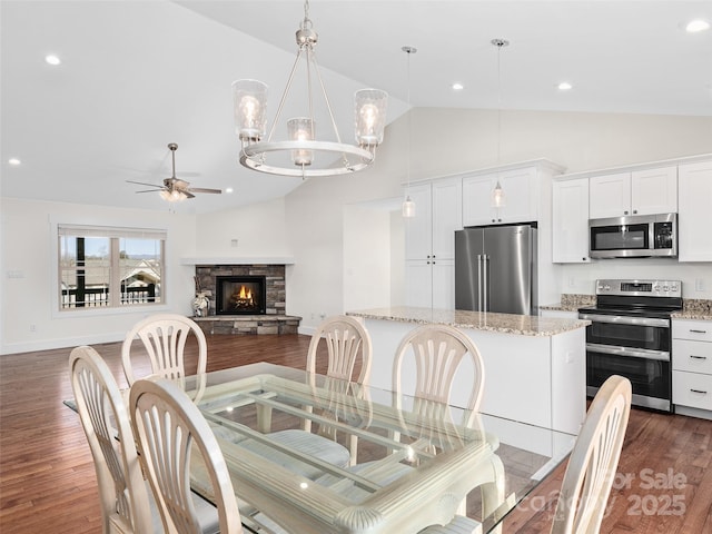 dining area featuring vaulted ceiling, dark hardwood / wood-style flooring, ceiling fan with notable chandelier, and a stone fireplace