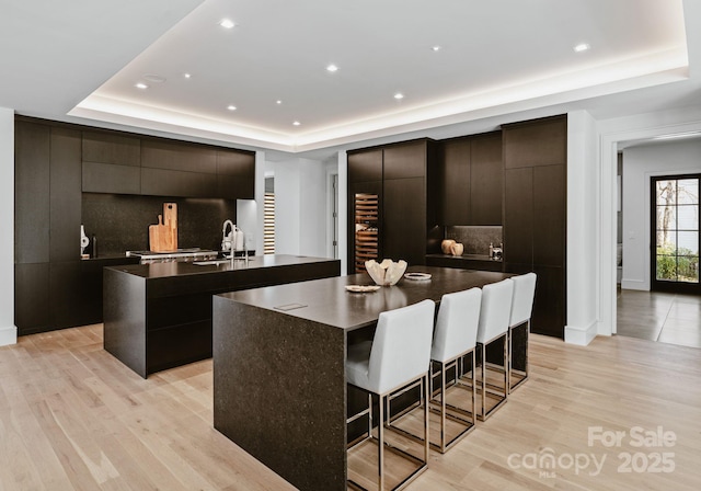 kitchen with a raised ceiling, an island with sink, dark countertops, light wood-style floors, and a kitchen bar