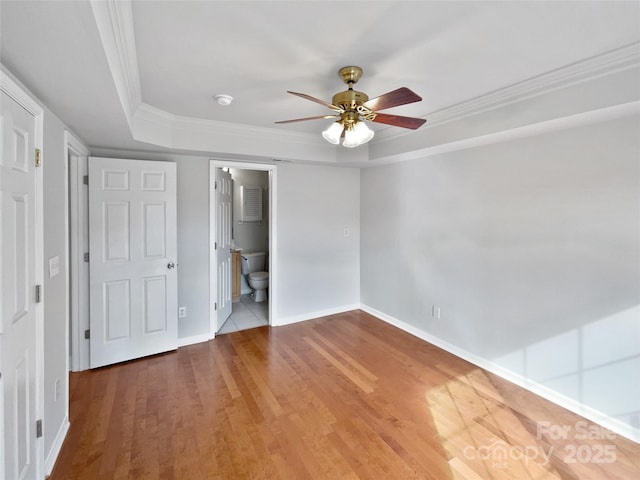 unfurnished bedroom featuring ceiling fan, hardwood / wood-style floors, ensuite bath, a tray ceiling, and ornamental molding