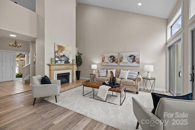 living area featuring a towering ceiling, a tiled fireplace, an inviting chandelier, light wood-style floors, and baseboards