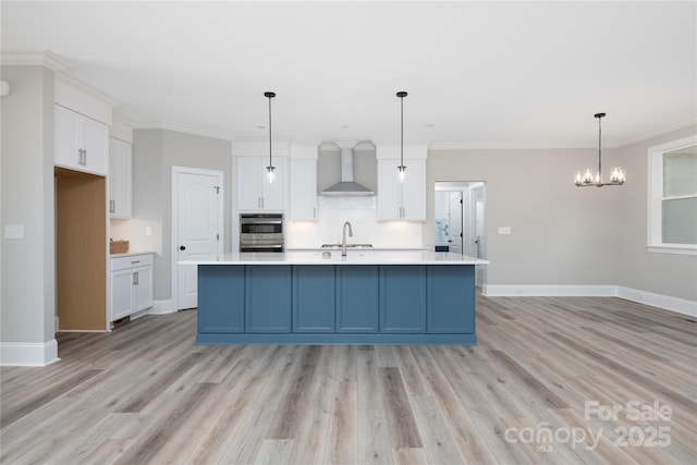 kitchen with white cabinetry, ornamental molding, pendant lighting, a kitchen island with sink, and wall chimney range hood