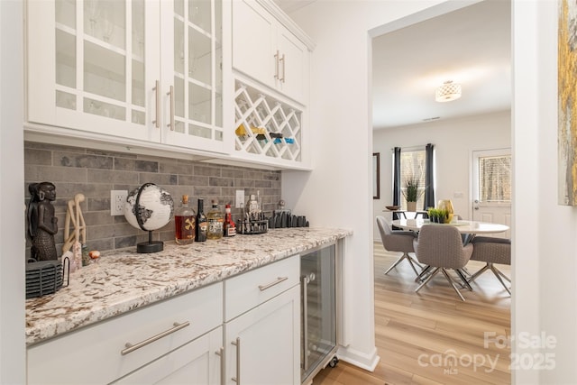 bar with decorative backsplash, beverage cooler, white cabinets, light stone countertops, and light hardwood / wood-style flooring