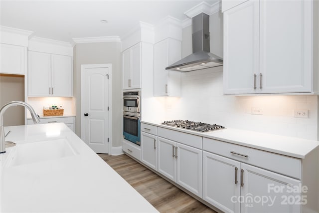 kitchen featuring appliances with stainless steel finishes, white cabinetry, wall chimney range hood, light hardwood / wood-style floors, and sink