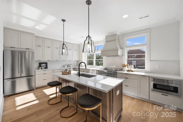 kitchen with custom exhaust hood, premium appliances, a center island with sink, decorative light fixtures, and sink