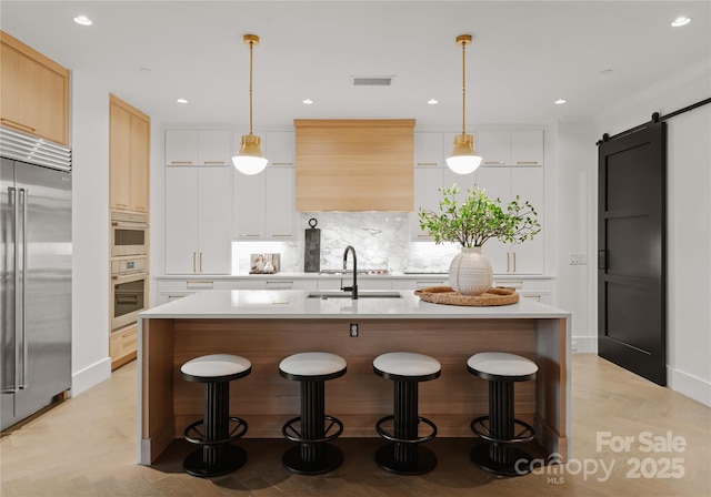 kitchen featuring a barn door, a kitchen island with sink, built in fridge, light parquet flooring, and sink