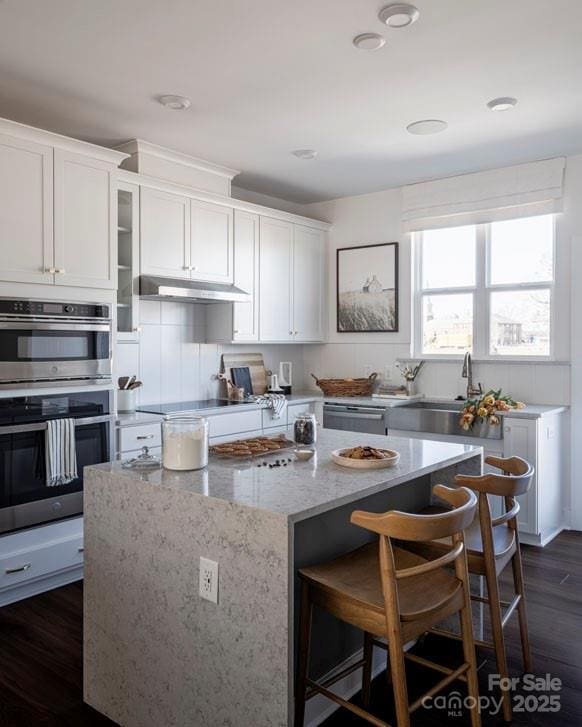 kitchen featuring dark wood-type flooring, a center island, light stone countertops, stainless steel appliances, and under cabinet range hood