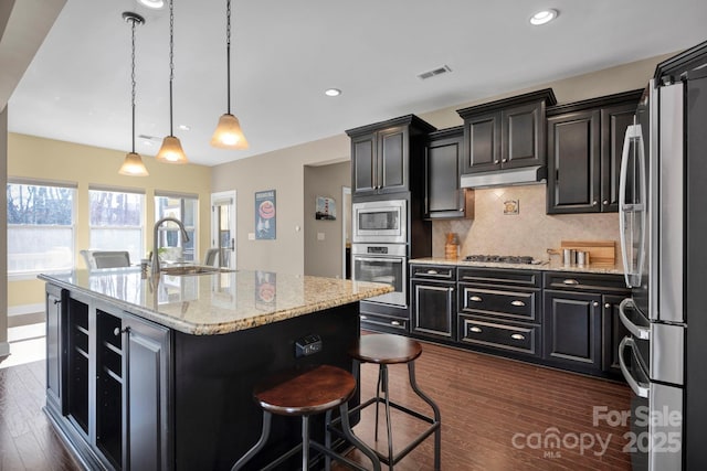 kitchen with light stone counters, stainless steel appliances, a center island with sink, and hanging light fixtures