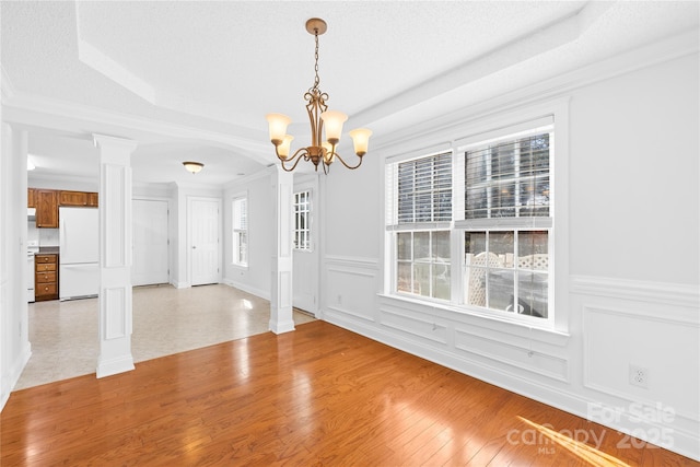 unfurnished dining area with ornate columns, a raised ceiling, light wood-type flooring, ornamental molding, and a chandelier