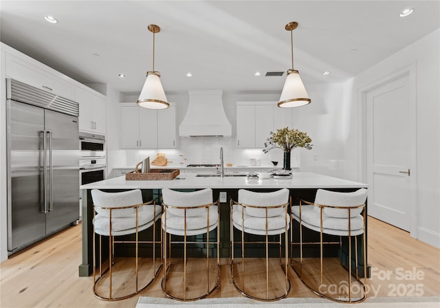 kitchen featuring premium range hood, stainless steel appliances, white cabinets, a center island with sink, and decorative light fixtures