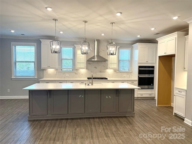 kitchen featuring an island with sink, dark hardwood / wood-style floors, hanging light fixtures, wall chimney range hood, and white cabinets