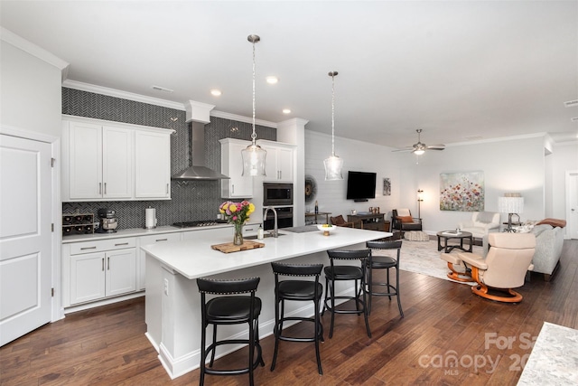 kitchen with white cabinetry, wall chimney exhaust hood, hanging light fixtures, and a kitchen island with sink