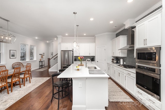 kitchen with decorative light fixtures, wall chimney range hood, stainless steel appliances, and a center island with sink