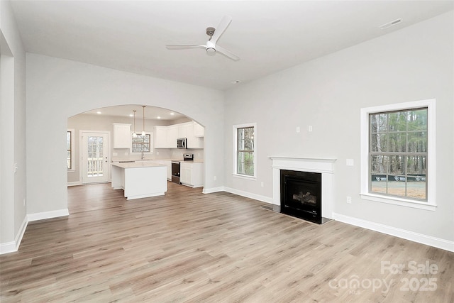 unfurnished living room featuring arched walkways, a fireplace, a ceiling fan, light wood-type flooring, and baseboards