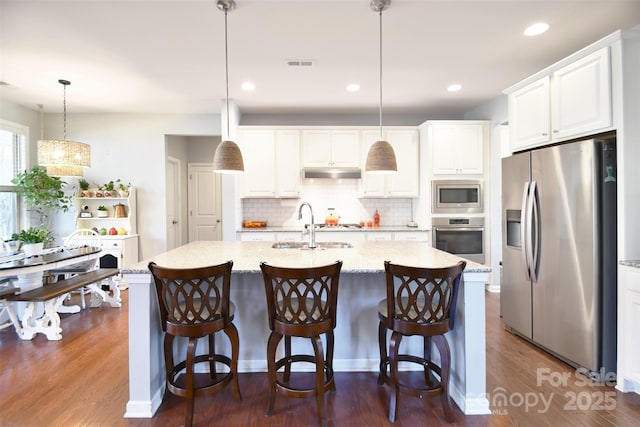 kitchen featuring pendant lighting, appliances with stainless steel finishes, light stone countertops, an island with sink, and white cabinets