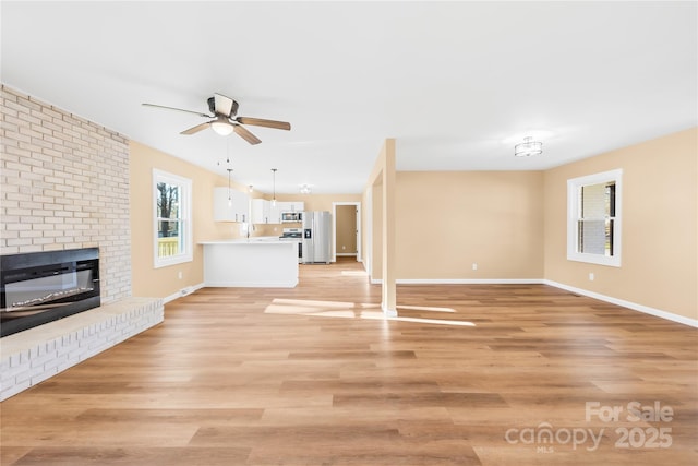 unfurnished living room with ceiling fan, a brick fireplace, baseboards, and light wood-style floors