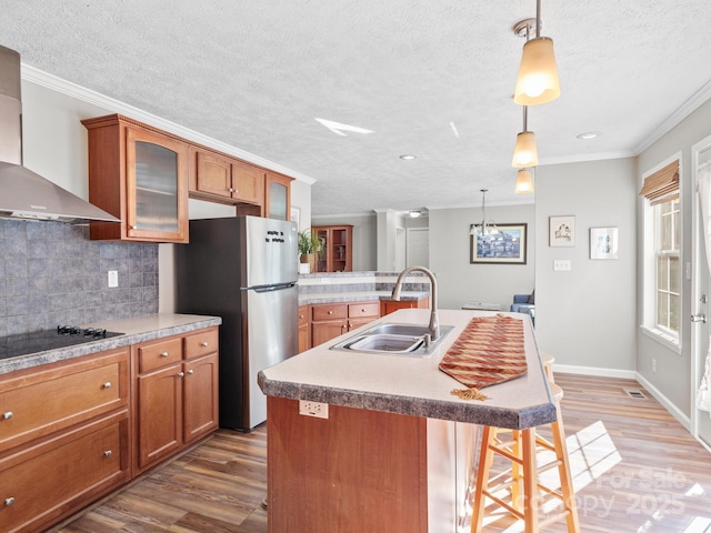 kitchen with a center island with sink, black electric cooktop, ornamental molding, wall chimney exhaust hood, and sink