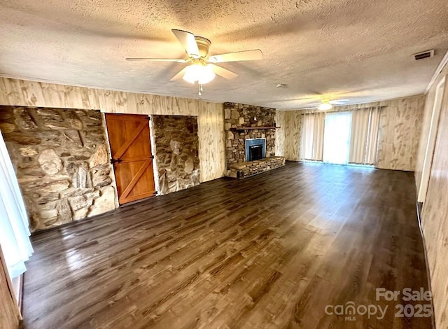 unfurnished living room with a textured ceiling, a stone fireplace, visible vents, a ceiling fan, and dark wood finished floors