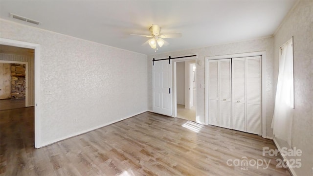 unfurnished bedroom featuring a barn door, light wood-style flooring, visible vents, a ceiling fan, and a closet