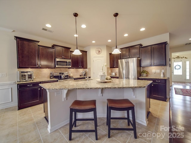 kitchen featuring appliances with stainless steel finishes, a breakfast bar area, hanging light fixtures, light stone countertops, and a center island with sink