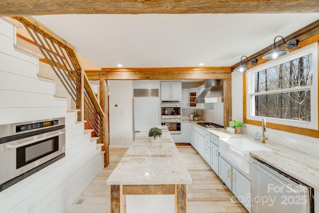 kitchen with sink, appliances with stainless steel finishes, a center island, light stone counters, and wall chimney exhaust hood