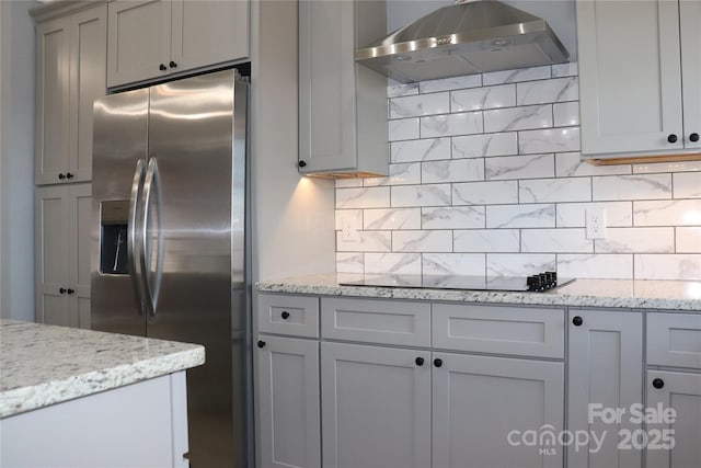 kitchen with wall chimney exhaust hood, light stone counters, stainless steel fridge with ice dispenser, black electric cooktop, and decorative backsplash