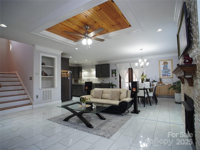 living room featuring ceiling fan with notable chandelier, built in shelves, a stone fireplace, ornamental molding, and wooden ceiling