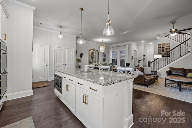 kitchen with white cabinetry, a center island with sink, stainless steel microwave, hanging light fixtures, and light stone counters