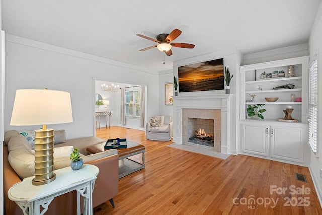 living room featuring a tile fireplace, light hardwood / wood-style floors, ornamental molding, built in shelves, and ceiling fan with notable chandelier