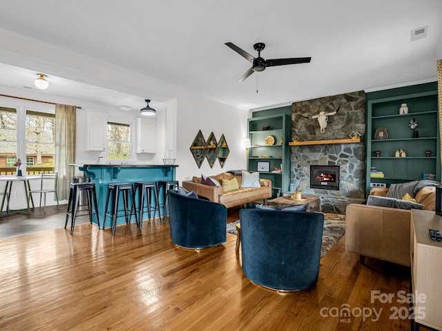 living room with built in shelves, a fireplace, ceiling fan, and light wood-type flooring