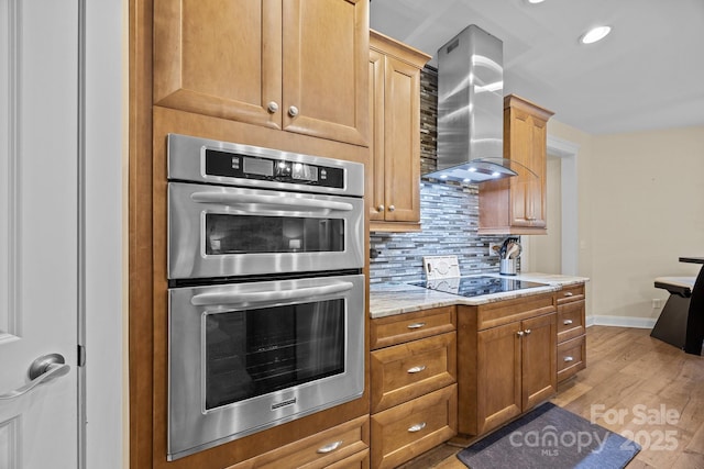 kitchen featuring backsplash, wall chimney range hood, black electric stovetop, stainless steel double oven, and light stone counters